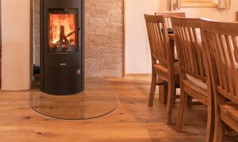 A cozy living space with a modern wood stove and a wooden dining table. The room features warm wood tones and plenty of light.