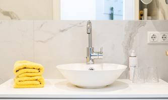A modern bathroom with a white sink and elegant mirror lighting. Stacks of yellow towels are neatly arranged on the surface.