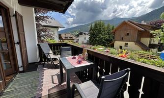 A cozy balcony with a table and two chairs. In the background, mountains and houses can be seen, while the clouds drift by in the sky.