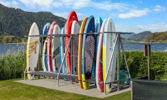 A row of stand-up paddle boards is neatly arranged on a rack by the water. In the background, green mountains and a clear blue sky can be seen.