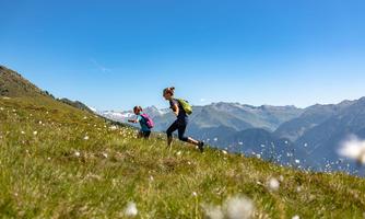 Two girls are running in a blooming meadow in the mountains. In the background, majestic mountains and a bright blue sky can be seen.