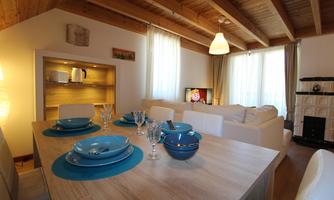 A cozy living room with wooden beams and large windows. The dining table is beautifully set with blue plates and glasses.