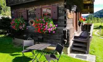 A cozy wooden cabin with colorful flowers at the windows. In front of the cabin, there is a small table and chairs on green grass.