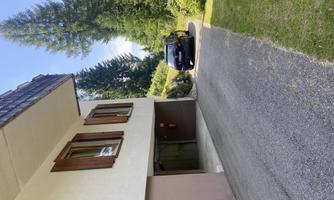 A building with wooden shutters and a driveway. Next to the building, there is a black car on a paved road.