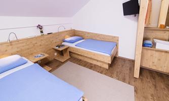 A bright room with two single beds and a modern wood design. In the background, a television and a small washing area are visible.