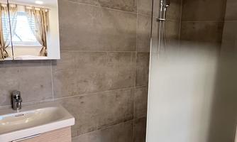 A modern bathroom with a glass shower and a simple sink. The walls are in neutral tones, and a window provides natural light.