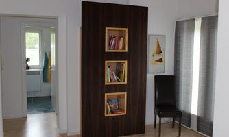 A modern room with a bookshelf that is integrated into a wall. In the background, a chair and a window are visible.