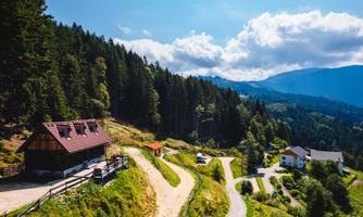 A picturesque landscape with a small wooden house, surrounded by green forests and mountains. The sky is clear with scattered clouds.