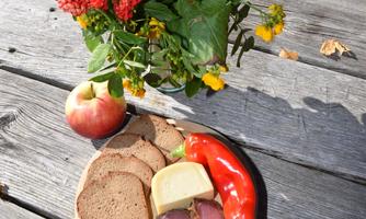 A wooden board with fresh bread, cheese, sausage, and a bell pepper, along with an apple. In the background, there is a vase with colorful flowers.