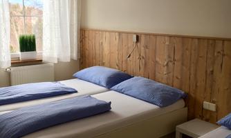 A bright bedroom with two beds and blue bedding. The wall is clad in wood and a window provides plenty of daylight.
