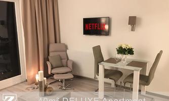 A modern apartment with a cozy armchair, a dining table for two, and a television displaying Netflix. The decor is bright and inviting.