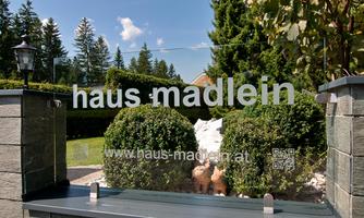 A well-maintained garden with round hedges and a clear blue sky. In the foreground stands a modern sign with the inscription "haus madlein".