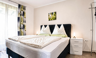 A bright bedroom with a large bed and several cushions. On the wall hangs a small picture and there is a bedside table with a lamp.