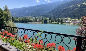 A peaceful lake surrounded by green mountains and trees. In the foreground, red flowers bloom on a terrace.