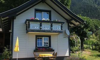 A pretty house with a balcony and colorful flowers. The garden is green and well-maintained, surrounded by mountains.