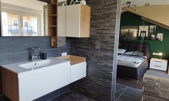 A modern bathroom with an elegant sink line and dark tiles. In the background, there is a bedroom with a cozy bed.