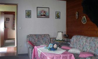 A cozy living room with a floral sofa and pastel-colored tablecloths. Pictures are hanging on the wall, and there is a lamp on the table.