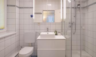 A modern bathroom with white tiles and clean lines. It features a shower, a sink, and a toilet.