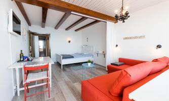 A modern room with a cozy red sofa and a double bed. The room has bright walls and large windows that provide plenty of light.