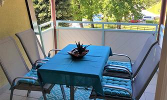 A cozy balcony with a table and chairs. The table is covered with a blue tablecloth and a decorative plant, while green trees are visible in the background.