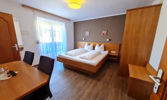 A bright bedroom with a double bed and two nightstands. A table with chairs is in the foreground and large windows provide plenty of daylight.