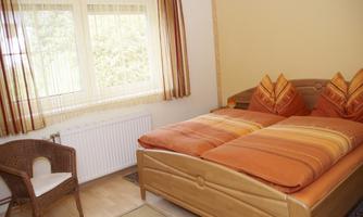A cozy bedroom with a double bed and orange pillows. Next to the bed is a chair, and large windows let in plenty of light.