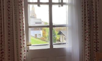 A window with white curtains that looks out onto a peaceful landscape. In the background, a church tower and other buildings are visible.