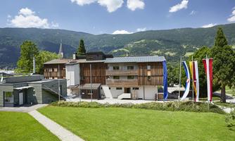 A modern building with a wooden facade in a green environment. In the background, hills and a blue sky with some clouds are visible.