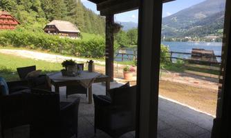 A cozy terrace with garden furniture and a view of a lake. In the background, green hills and rustic houses can be seen.