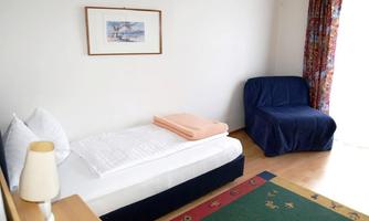 A simple bedroom with a single bed and a blue armchair. The room has a bright, cheerful look with a carpet and a window with curtains.