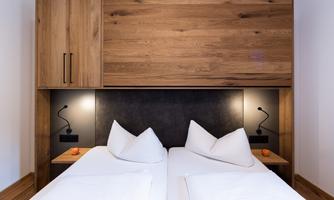 A modern bedroom with two single beds and a wood finish. Two bedside lamps and storage space are located on the wall.