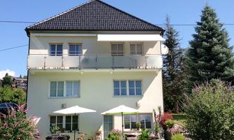 A modern house with two floors and a balcony. The garden is green, with plants and sun umbrellas.