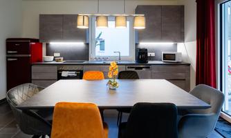 A modern kitchen with a large dining table and colorful chairs. The walls are kept neutral, and there is decorative lighting above the table.