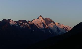 A majestic mountain with a snow-capped peak in the early morning light. The surrounding mountains are wrapped in gentle shadows.