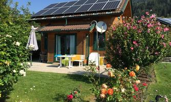 A cozy wooden house with solar panels on the roof. The garden is blooming with colorful roses and an outdoor seating area.