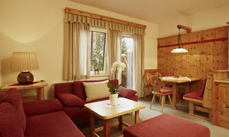A cozy living space with red sofas and a wooden table. Bright windows and a warm atmosphere create an inviting environment.