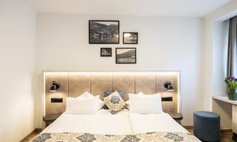 A modern bedroom with a large double bed and elegant decoration. Photos hang on the wall, and a small table stands next to a window with flowers.
