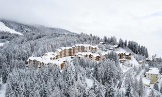 A snowy mountain landscape with a picturesque village, surrounded by tall, green fir trees. The sky is cloudy, giving the scene a tranquil atmosphere.