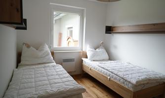 A simple bedroom with two single beds and a window. The room features a modern, minimalist style with wooden details.
