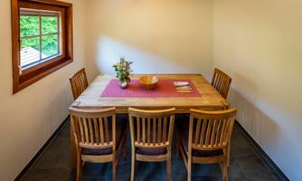 A simply furnished dining area with a wooden table and five chairs. A red-brown tablecloth and a vase of flowers are on the table.
