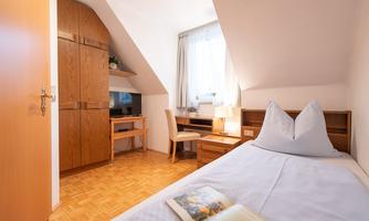 A cozy bedroom with a single bed, a desk, and a television. The light shines through the window, creating an inviting atmosphere.