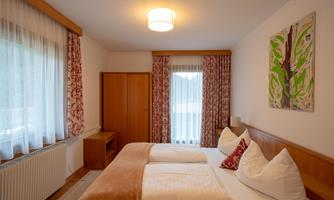A cozy bedroom with two beds and large windows. The walls are light, and there are decorative curtains as well as a piece of artwork on the wall.