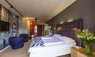 A modern hotel room with a comfortable double bed and stylish furnishings. The walls are in neutral colors, and there is a small sitting area as well as a floral decoration.