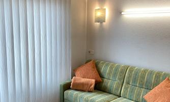 A cozy living room with a green couch and two orange cushions. A wooden table stands in front of the couch, and large windows with white curtains let in plenty of light.