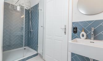 A modern bathroom with a shower and teal wall tiles. An elegant sink and a round mirror are also present.