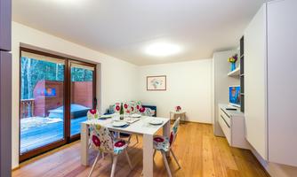 A bright and modern dining room with a white table and colorful chairs. Large windows provide a view of a wooden terrace and the surrounding nature.