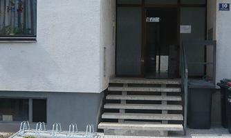The entrance area of a building with steps and an entrance door. A sign with the name of the institution is mounted above the door.
