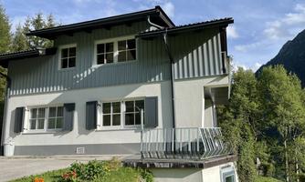 A modern house with a beautiful balcony and large windows. Surrounded by green trees and a clear blue sky.
