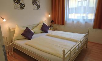 A cozy bedroom with a double bed and two cushions in purple. Large windows let in plenty of light, and the walls are decorated with floral pictures.