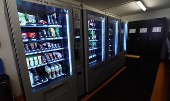 Three modern vending machines are located in a room. They offer a variety of snacks and drinks.